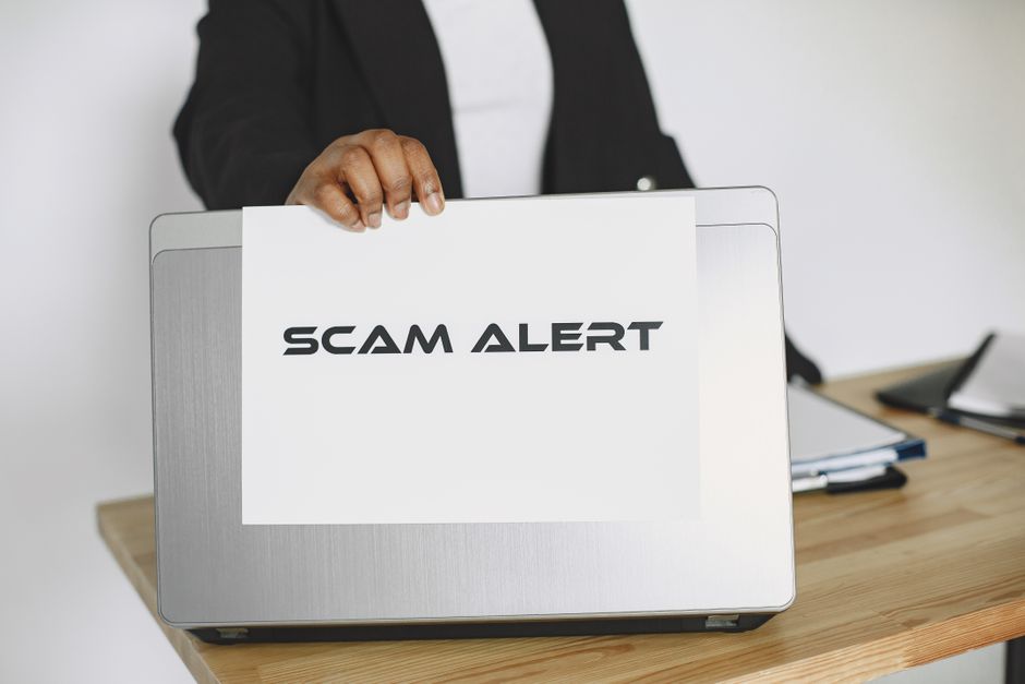 Osoba trzymająca kartkę z napisem SCAM ALERT na tle laptopa.