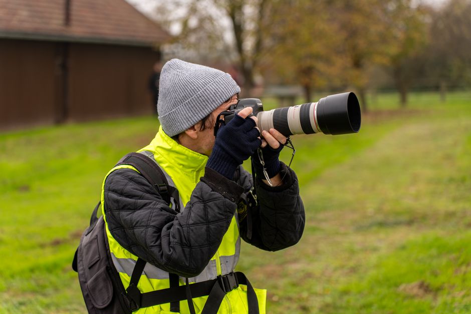 Fotograf w szarej czapce i kamizelce odblaskowej używający aparatu z teleobiektywem.