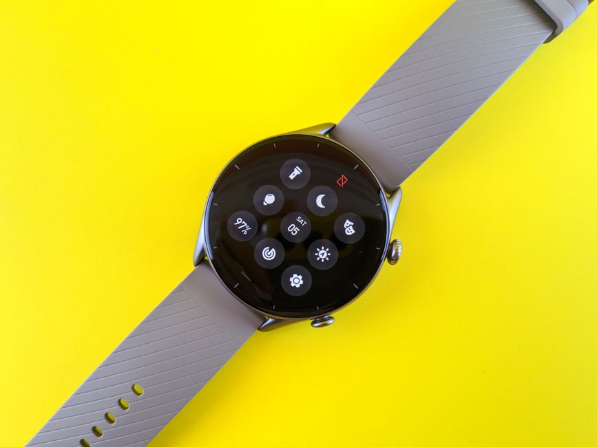 Smartwatch z szarym paskiem wyświetlający menu ustawień na żółtym tle